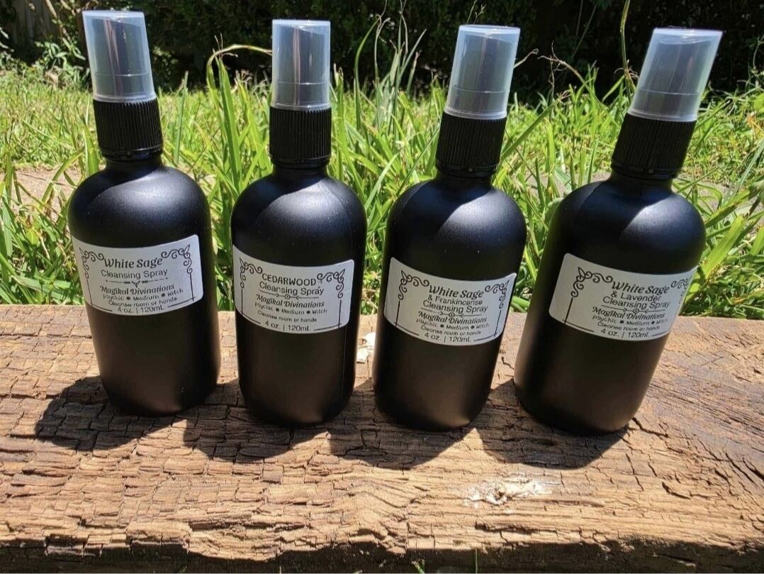 Four black Magikal Divinations cleansing spray bottles lined up on a weathered wooden plank in the sun, labeled White Sage, Cedarwood, White Sage & Frankincense, and White Sage & Lavender